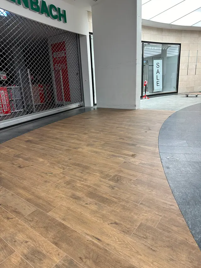 Einkaufszentrum, Ladeneingang: Holzoptik-Planken vor dem Ladeneingang, gebogener Übergang zum Mallboden