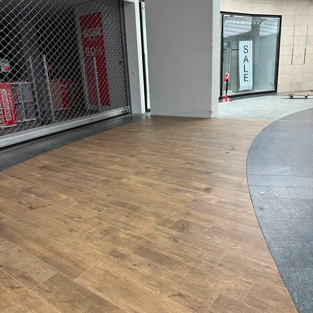 Einkaufszentrum, Ladeneingang: Holzoptik-Planken vor dem Ladeneingang, gebogener Übergang zum Mallboden
